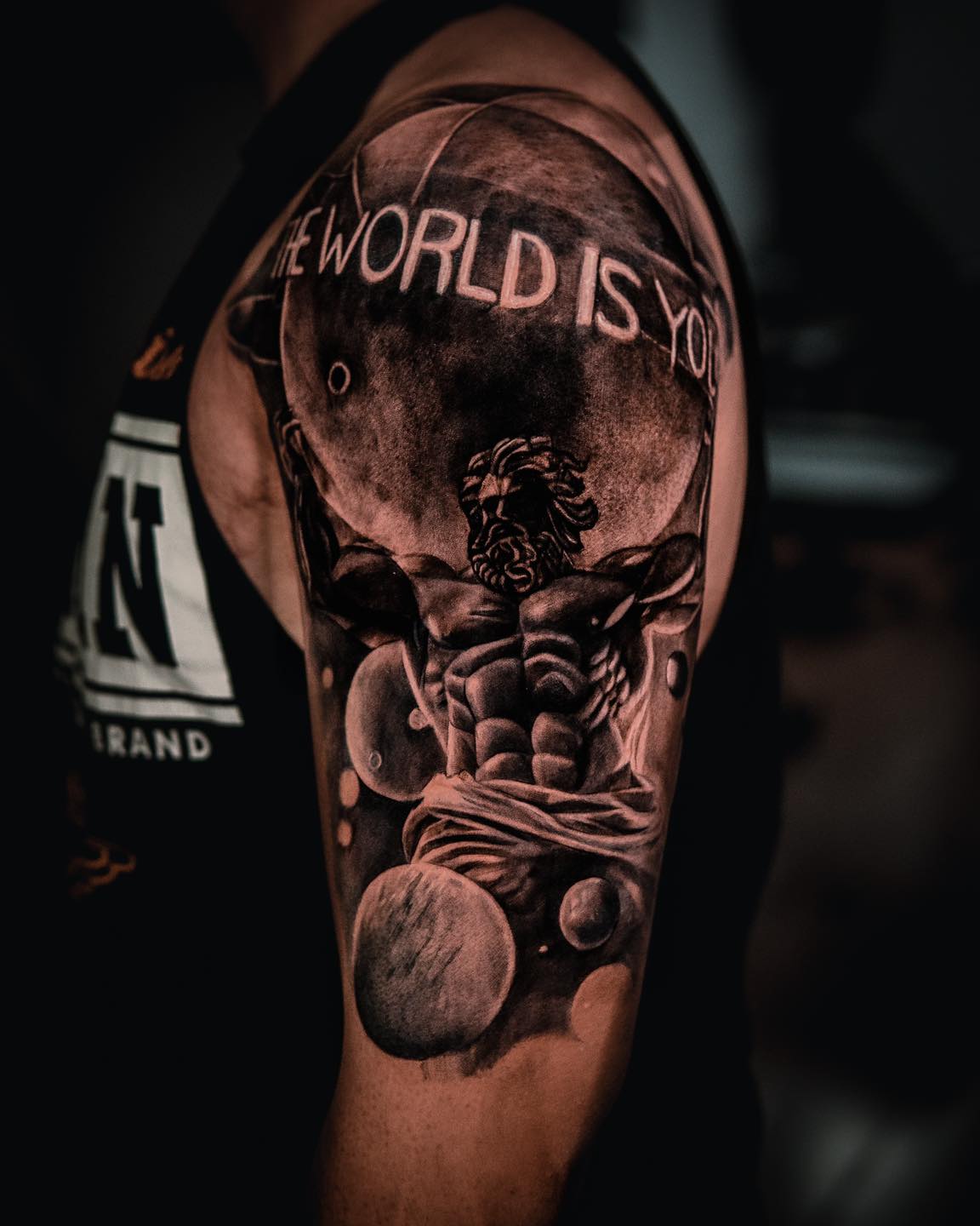 Tatuaje realista de Atlas cargando el mundo con lettering The World Is Yours, brazo completo